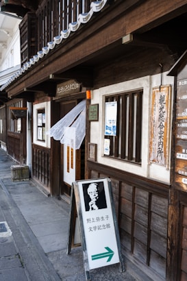 A traditional wooden building with sliding windows and lanterns hanging at the entrance. A sandwich board on the sidewalk features a black and white image of a person and Japanese text. Wooden signs with Japanese writing hang next to the entrance, and a cloth drapes across the doorway.