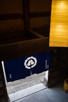 A traditional interior space with a blue fabric divider featuring a white flower-like emblem. A large wooden beam crosses above the divider, and below, there is an open doorway leading outside to a stone pathway. Part of a softly lit yellow paper lantern is visible in the upper right corner.