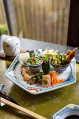 A beautifully arranged plate features various small dishes, including fried tempura, pickled vegetables, and garnished bowls containing rice and green onions. The intricate design on the plate complements the carefully presented food. A ceramic lidded bowl and chopsticks rest nearby, creating an inviting and authentic dining experience.