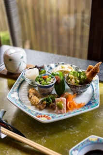 A beautifully arranged plate features various small dishes, including fried tempura, pickled vegetables, and garnished bowls containing rice and green onions. The intricate design on the plate complements the carefully presented food. A ceramic lidded bowl and chopsticks rest nearby, creating an inviting and authentic dining experience.