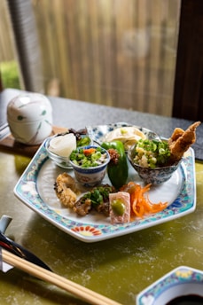 A beautifully arranged plate features various small dishes, including fried tempura, pickled vegetables, and garnished bowls containing rice and green onions. The intricate design on the plate complements the carefully presented food. A ceramic lidded bowl and chopsticks rest nearby, creating an inviting and authentic dining experience.