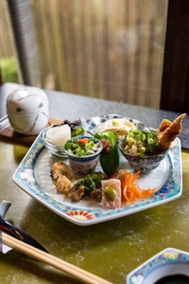 A beautifully arranged plate features various small dishes, including fried tempura, pickled vegetables, and garnished bowls containing rice and green onions. The intricate design on the plate complements the carefully presented food. A ceramic lidded bowl and chopsticks rest nearby, creating an inviting and authentic dining experience.