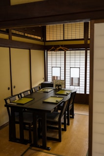 A traditional Japanese room featuring a dark wooden table set with green placemats, arranged on a tatami mat floor. The room is enclosed with shoji screens, creating a serene and minimalist atmosphere. Two wooden hangers are visible on the wall, adding to the rustic and organized aesthetic.