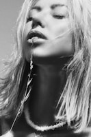 A woman with closed eyes holds a delicate plant stem near her lips, creating a serene and contemplative mood. Her hair is slightly tousled, and she wears a simple necklace.