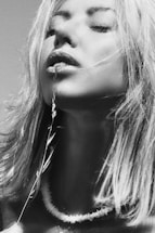 A woman with closed eyes holds a delicate plant stem near her lips, creating a serene and contemplative mood. Her hair is slightly tousled, and she wears a simple necklace.