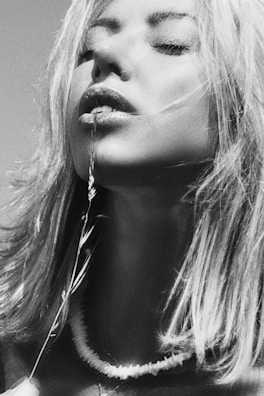A woman with closed eyes holds a delicate plant stem near her lips, creating a serene and contemplative mood. Her hair is slightly tousled, and she wears a simple necklace.