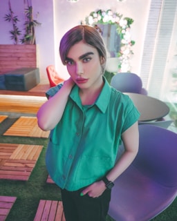 A person wearing a green shirt stands indoors, surrounded by colorful and modern furniture. The setting includes a wooden floor, a decorative wall with plants, and a contemporary round table and chairs. Soft, colorful lighting enhances the atmosphere.