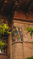 A section of an architectural structure features intricate colored glass in a geometric pattern set into a brick wall. Wooden elements frame the glasswork, and green foliage partially obscures the view, suggesting an outdoor setting.