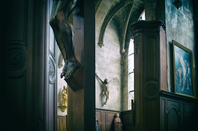 The interior of a church with a prominent crucifix and several religious statues. The walls and columns feature detailed carvings, and there is a framed religious painting on the wall. Soft light filters through stained glass windows, creating a serene and reverent atmosphere.