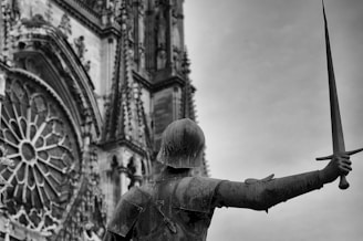 A historical statue of a knight in armor is seen holding a sword aloft in front of a Gothic cathedral facade. The intricate architecture, including a round rose window and detailed stonework, creates a dramatic backdrop.