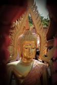 A golden Buddha statue is adorned with intricate designs, featuring a serene expression and a ruby-like gem on its forehead. The statue is surrounded by people, with ornate decorations in the background, possibly part of a religious ceremony or temple setting.