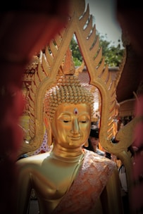 A golden Buddha statue is adorned with intricate designs, featuring a serene expression and a ruby-like gem on its forehead. The statue is surrounded by people, with ornate decorations in the background, possibly part of a religious ceremony or temple setting.