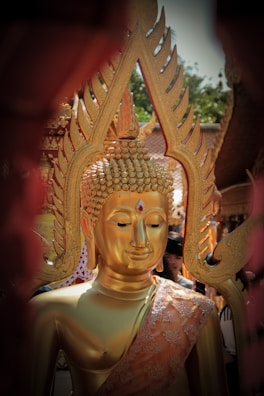 A golden Buddha statue is adorned with intricate designs, featuring a serene expression and a ruby-like gem on its forehead. The statue is surrounded by people, with ornate decorations in the background, possibly part of a religious ceremony or temple setting.