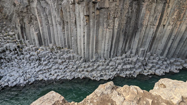 A dramatic view of towering vertical basalt columns, resembling a natural fortress wall, with a river flowing at its base. These columns show the unique geometric formation typical of volcanic activity, with sharp, angular lines and various shades of gray.