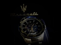 A luxury wristwatch with a detailed open-worked dial showcasing its intricate mechanical movements. The watch is silver with a blue and black color theme and features bold numerals on the bezel. Behind the watch, there is a blurred Maserati logo, suggestive of the brand's emblematic style.
