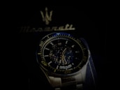 A luxury wristwatch with a detailed open-worked dial showcasing its intricate mechanical movements. The watch is silver with a blue and black color theme and features bold numerals on the bezel. Behind the watch, there is a blurred Maserati logo, suggestive of the brand's emblematic style.