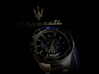 A luxury wristwatch with a detailed open-worked dial showcasing its intricate mechanical movements. The watch is silver with a blue and black color theme and features bold numerals on the bezel. Behind the watch, there is a blurred Maserati logo, suggestive of the brand's emblematic style.