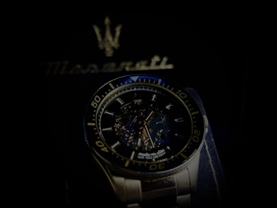 A luxury wristwatch with a detailed open-worked dial showcasing its intricate mechanical movements. The watch is silver with a blue and black color theme and features bold numerals on the bezel. Behind the watch, there is a blurred Maserati logo, suggestive of the brand's emblematic style.