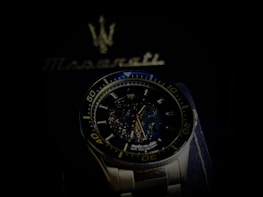 A luxury wristwatch with a detailed open-worked dial showcasing its intricate mechanical movements. The watch is silver with a blue and black color theme and features bold numerals on the bezel. Behind the watch, there is a blurred Maserati logo, suggestive of the brand's emblematic style.