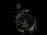A luxury wristwatch with a detailed open-worked dial showcasing its intricate mechanical movements. The watch is silver with a blue and black color theme and features bold numerals on the bezel. Behind the watch, there is a blurred Maserati logo, suggestive of the brand's emblematic style.
