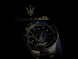 A luxury wristwatch with a detailed open-worked dial showcasing its intricate mechanical movements. The watch is silver with a blue and black color theme and features bold numerals on the bezel. Behind the watch, there is a blurred Maserati logo, suggestive of the brand's emblematic style.