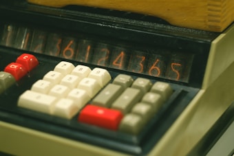 A close-up view of an old-fashioned calculator displaying numbers on a nixie tube display. The calculator has a number pad with red, white, and grey keys, including function keys labeled 'Ck' and 'C'. The device appears to be made of beige plastic material with a slightly textured finish.