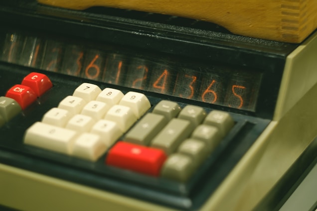 A close-up view of an old-fashioned calculator displaying numbers on a nixie tube display. The calculator has a number pad with red, white, and grey keys, including function keys labeled 'Ck' and 'C'. The device appears to be made of beige plastic material with a slightly textured finish.