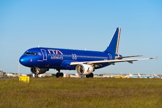An airplane with a blue fuselage is on the runway, featuring the logo of ITA Airways on its side. The aircraft is positioned on a grassy field with a clear blue sky in the background, suggesting it's prepared for takeoff or has just landed.