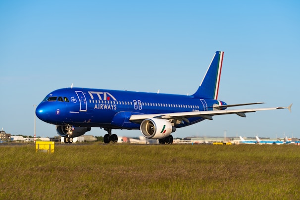 An airplane with a blue fuselage is on the runway, featuring the logo of ITA Airways on its side. The aircraft is positioned on a grassy field with a clear blue sky in the background, suggesting it's prepared for takeoff or has just landed.