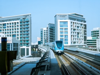 A modern and sleek metro train travels along elevated tracks surrounded by high-rise buildings with glass facades. The area is urban and well-developed, indicating a bustling city environment.