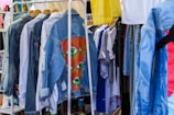 A collection of clothing items hanging on a rack, featuring denim jackets in the foreground with one jacket displaying colorful eye-themed artwork. Various other garments, including t-shirts and pants, are visible in the background, creating a vibrant and eclectic atmosphere.