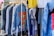 A collection of clothing items hanging on a rack, featuring denim jackets in the foreground with one jacket displaying colorful eye-themed artwork. Various other garments, including t-shirts and pants, are visible in the background, creating a vibrant and eclectic atmosphere.