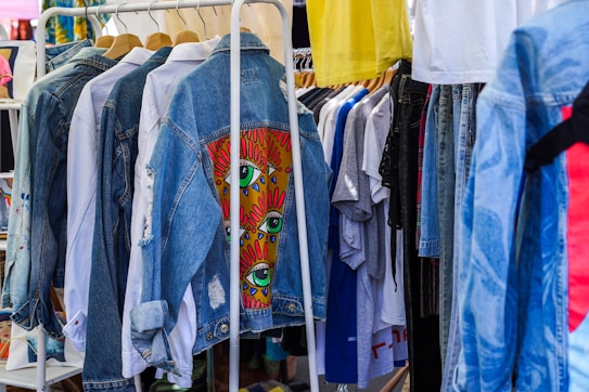 A collection of clothing items hanging on a rack, featuring denim jackets in the foreground with one jacket displaying colorful eye-themed artwork. Various other garments, including t-shirts and pants, are visible in the background, creating a vibrant and eclectic atmosphere.