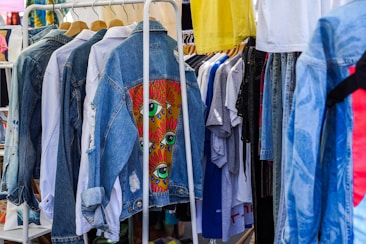 A collection of clothing items hanging on a rack, featuring denim jackets in the foreground with one jacket displaying colorful eye-themed artwork. Various other garments, including t-shirts and pants, are visible in the background, creating a vibrant and eclectic atmosphere.