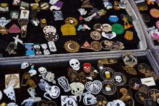 A collection of various decorative enamel pins displayed on black fabric. The pins feature diverse designs, including skulls, hearts, sun motifs, mystical symbols, and abstract patterns in an array of vibrant colors such as red, gold, blue, and black.