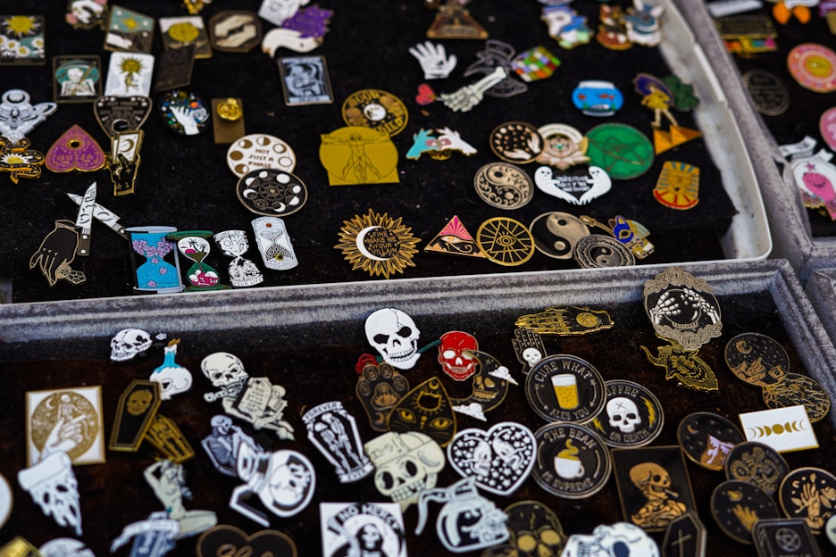 A collection of various decorative enamel pins displayed on black fabric. The pins feature diverse designs, including skulls, hearts, sun motifs, mystical symbols, and abstract patterns in an array of vibrant colors such as red, gold, blue, and black.