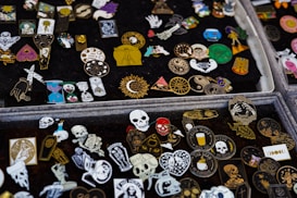A collection of various decorative enamel pins displayed on black fabric. The pins feature diverse designs, including skulls, hearts, sun motifs, mystical symbols, and abstract patterns in an array of vibrant colors such as red, gold, blue, and black.