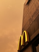 A concrete building facade with a prominent yellow letter 'M' extends upwards against a cloudy, sepia-toned sky.