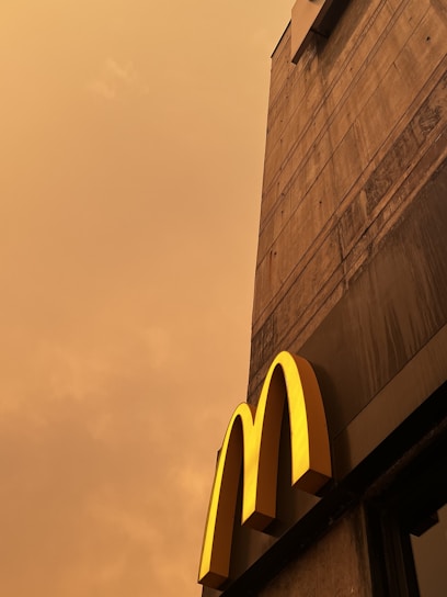 A concrete building facade with a prominent yellow letter 'M' extends upwards against a cloudy, sepia-toned sky.