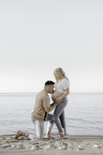 A couple is on a beach, with the man kneeling and gently holding the woman's pregnant belly as she stands, conveying tenderness and anticipation. The scene captures a serene and intimate moment with a calm sea and clear sky as the backdrop.
