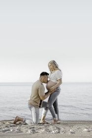 A couple is on a beach, with the man kneeling and gently holding the woman's pregnant belly as she stands, conveying tenderness and anticipation. The scene captures a serene and intimate moment with a calm sea and clear sky as the backdrop.