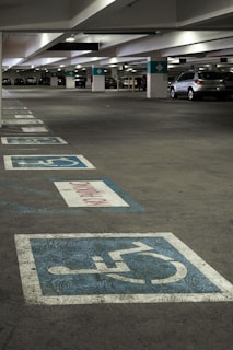 A multi-level parking garage featuring painted handicap parking symbols on the ground. Several cars are parked in the background, with the area dimly lit by overhead lights. Concrete pillars and signs denote parking sections.