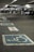 A multi-level parking garage featuring painted handicap parking symbols on the ground. Several cars are parked in the background, with the area dimly lit by overhead lights. Concrete pillars and signs denote parking sections.