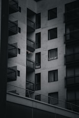 A modern apartment building with multiple balconies. The building is constructed with a minimalist design, featuring large glass windows and concrete walls. Some small lights or decorations are visible on one of the balconies.