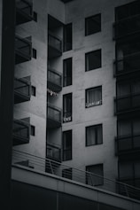 A modern apartment building with multiple balconies. The building is constructed with a minimalist design, featuring large glass windows and concrete walls. Some small lights or decorations are visible on one of the balconies.