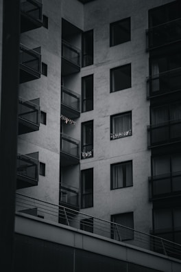 A modern apartment building with multiple balconies. The building is constructed with a minimalist design, featuring large glass windows and concrete walls. Some small lights or decorations are visible on one of the balconies.