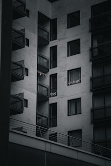 A modern apartment building with multiple balconies. The building is constructed with a minimalist design, featuring large glass windows and concrete walls. Some small lights or decorations are visible on one of the balconies.