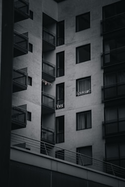 A modern apartment building with multiple balconies. The building is constructed with a minimalist design, featuring large glass windows and concrete walls. Some small lights or decorations are visible on one of the balconies.