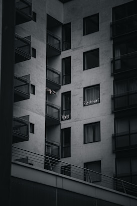 A modern apartment building with multiple balconies. The building is constructed with a minimalist design, featuring large glass windows and concrete walls. Some small lights or decorations are visible on one of the balconies.
