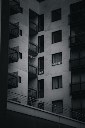 A modern apartment building with multiple balconies. The building is constructed with a minimalist design, featuring large glass windows and concrete walls. Some small lights or decorations are visible on one of the balconies.